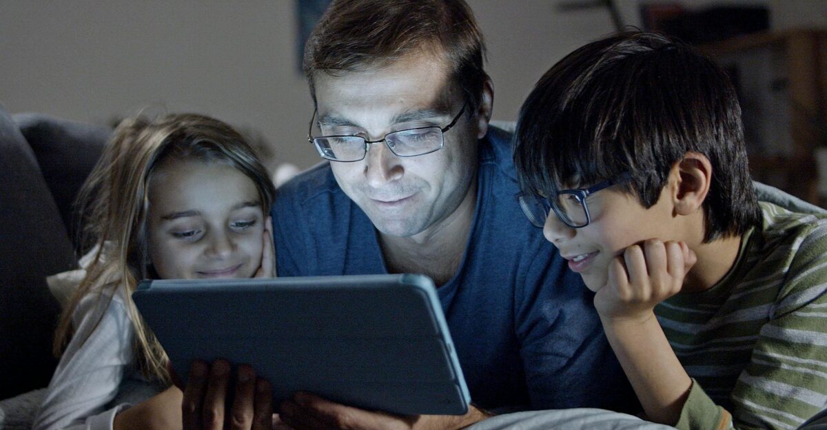A father and his children enjoy a story together on a tablet in a cozy indoor setting