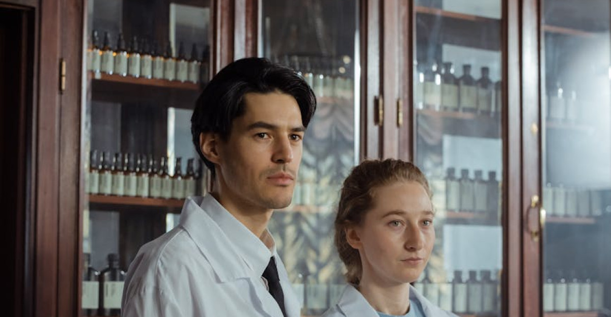 Two pharmacy employees in white coats standing by a wooden cabinet with shelves of bottles