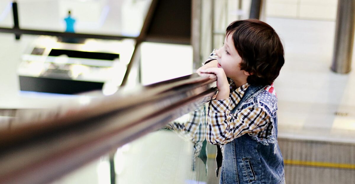 A young boy wearing a denim vest leans on the railing of an escalator inside a shopping mall