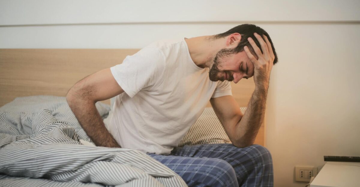 A young man in pajamas holding his head sitting on a bed appears to be experiencing a headache