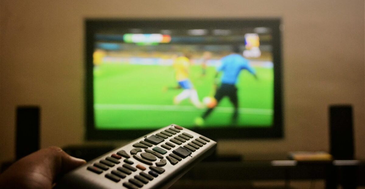 Close-up of a hand holding a remote control in front of a TV showing a football match