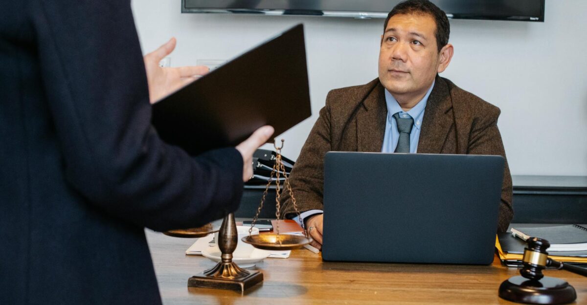 Crop unrecognizable employee representing new case details to concentrated middle aged ethnic lawyer sitting at table with laptop gavel and justice scales