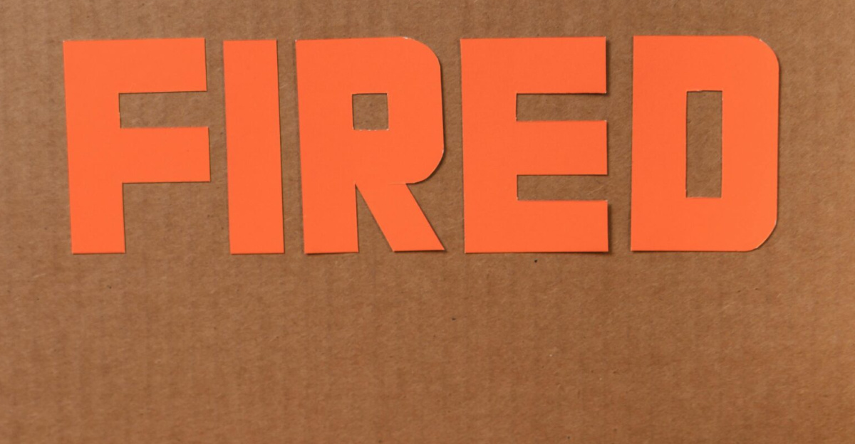 Close-up of a cardboard box labeled FIRED on a wooden table with crumpled papers.